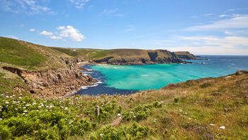 Nanjizal Colours This landscape photograph captures the vivid Nanjizal Colours along the coastline of Nanjizal Beach in Cornwall, England, United Kingdom. Taken during a summer afternoon, the image presents the turquoise sea stretching out into the Atlantic Ocean and bordered by rugged cliffs characteristic of the Cornish coast. Wildflowers and green vegetation cover the foreground, with rocky cliffs descending sharply into the clear waters below. The scenery highlights the natural beauty of Nanjizal Beach, with the cliffs framing the secluded cove and the bright summer sky overhead adding to the vibrant colours typical of this region.